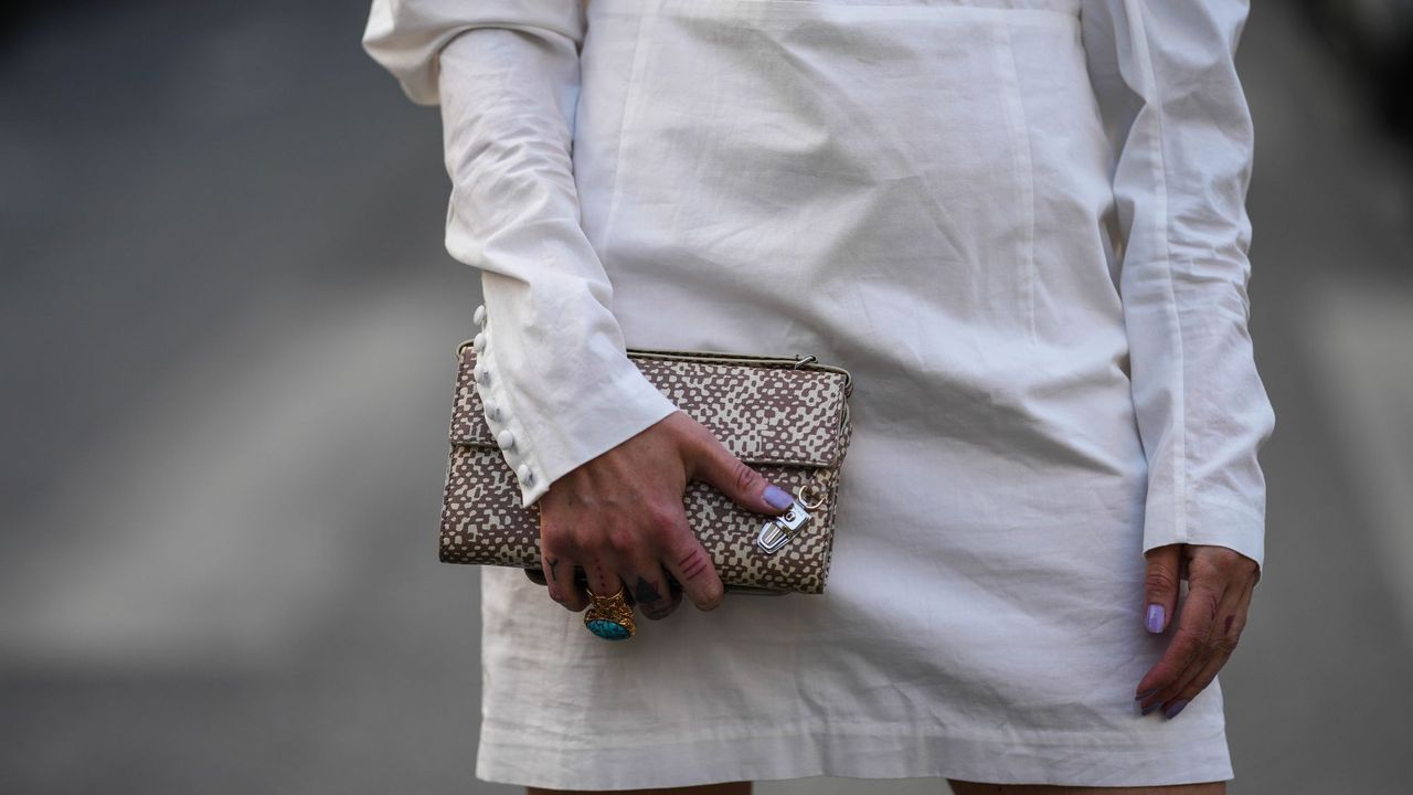 A crop of a woman wearing a white long-sleeved dress holding a snakeskin clutch bag and lavender nails