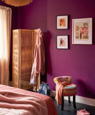 A deep purple bedroom with a corner of a pink bed, wall art on the walls, a wooden screen and hanging light, and a small patchwork chair at the foot of the bed with clothes on