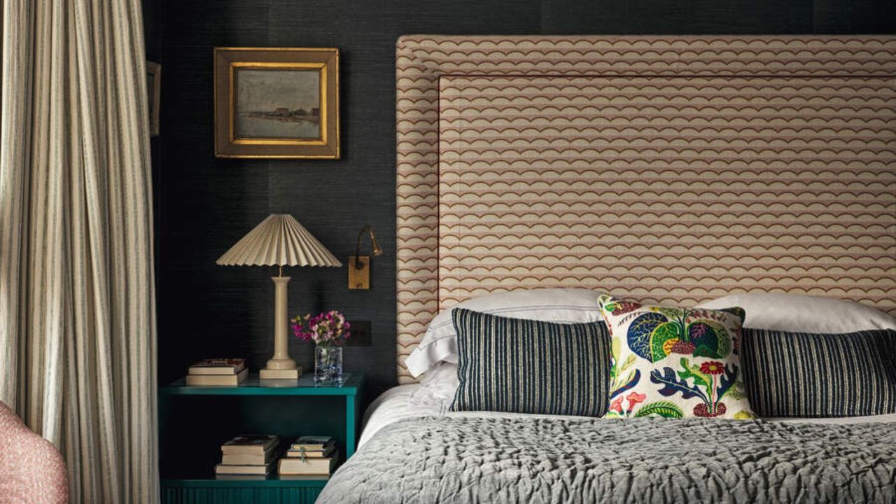 Gray bedding and blue and gray striped sheets on a bed with a tall brown headboard; a lamp on a blue nightstand against a gray wall.