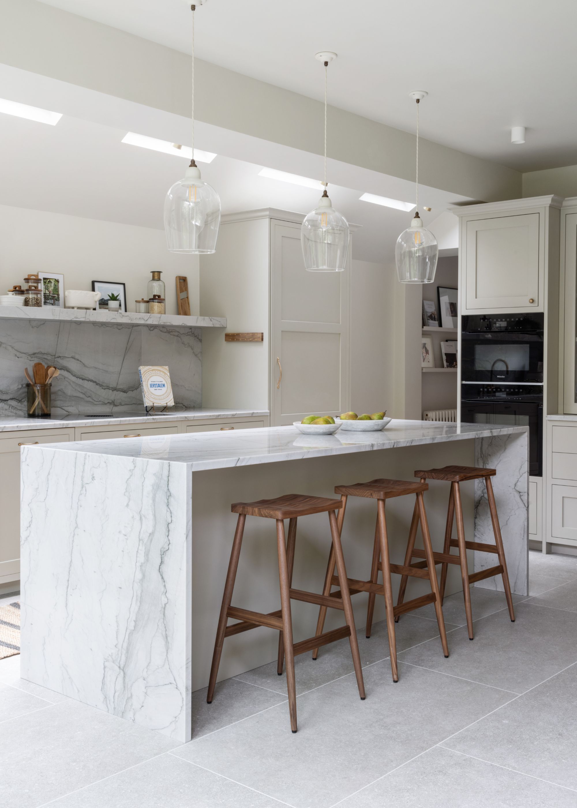 A kitchen with stone flooring and a stone backsplash. There is also a kitchen island with stools beside it, as well as three pendant lights hanging above
