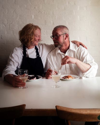 Female and male chef sat next to each other smiling at each other with food and drink on their table. 