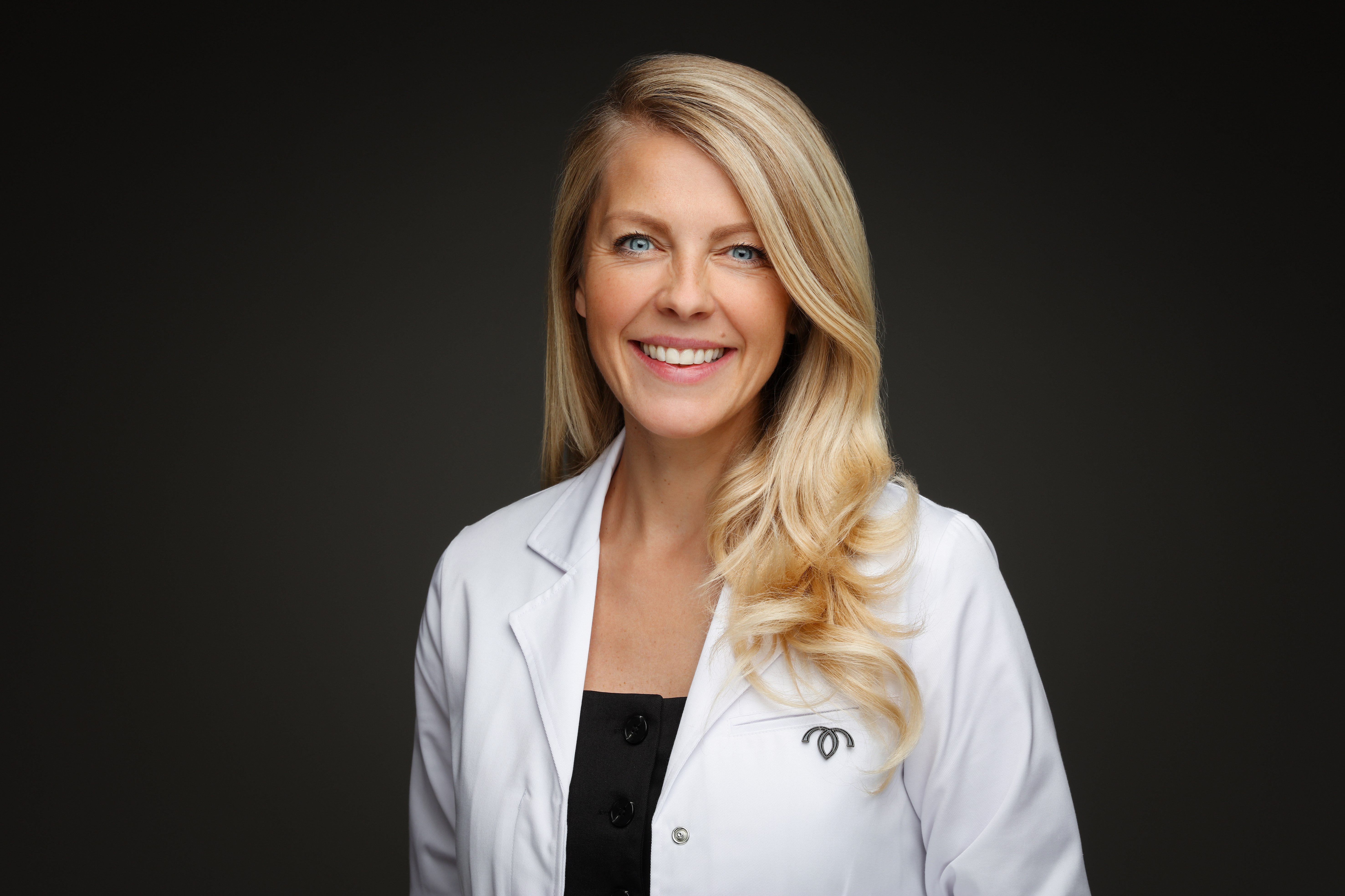 A headshot of a smiling blonde haired woman wearing a white lab coat with the Hertility logo