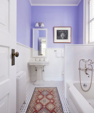 A bathroom with lilac painted walls above the dado rail, white tiles and bath, and red Persian-style rug
