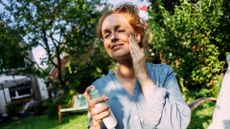 Woman putting after sun lotion on face, wearing shirt and standing in garden with tree behind her, after learning how to heal sunburn faster