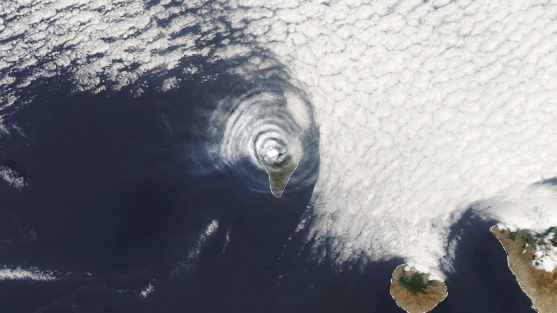 A satellite photo of an island covered with concentric cloud rings in the middle of the ocean