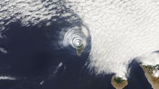 A satellite photo of an island covered with concentric cloud rings in the middle of the ocean