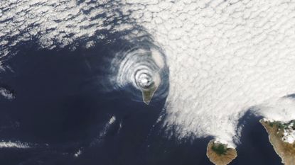 A satellite photo of an island covered with concentric cloud rings in the middle of the ocean
