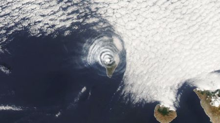 A satellite photo of an island covered with concentric cloud rings in the middle of the ocean