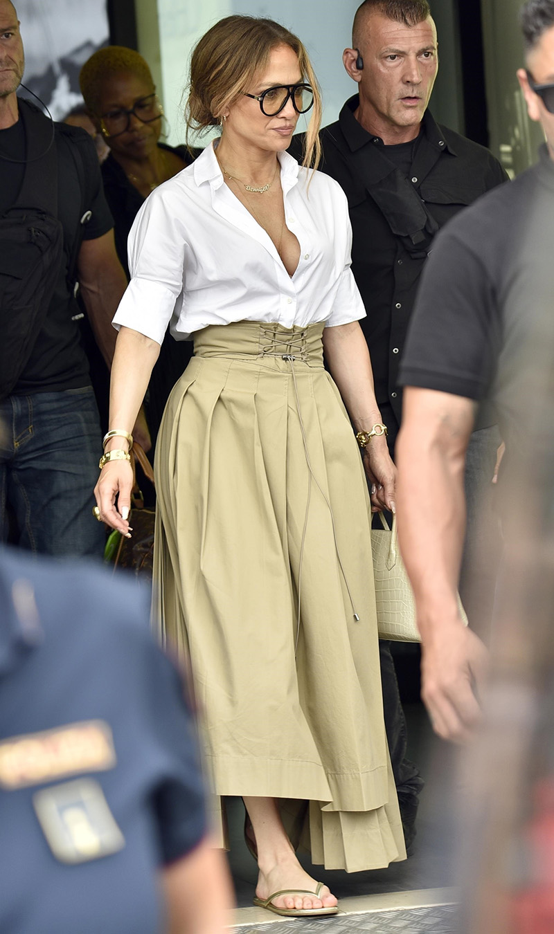 Jennifer Lopez wearing a white button-down, tan midi skirt, and flat leather sandals at the airport in Pisa, Italy.