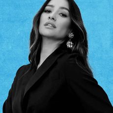 A black and white close up headshot of actress and entrepreneur Shay Mitchell, set on a blue background. 