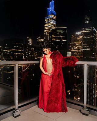 Cardi B wears a red gown with a red fur coat in front of the new york city skyline