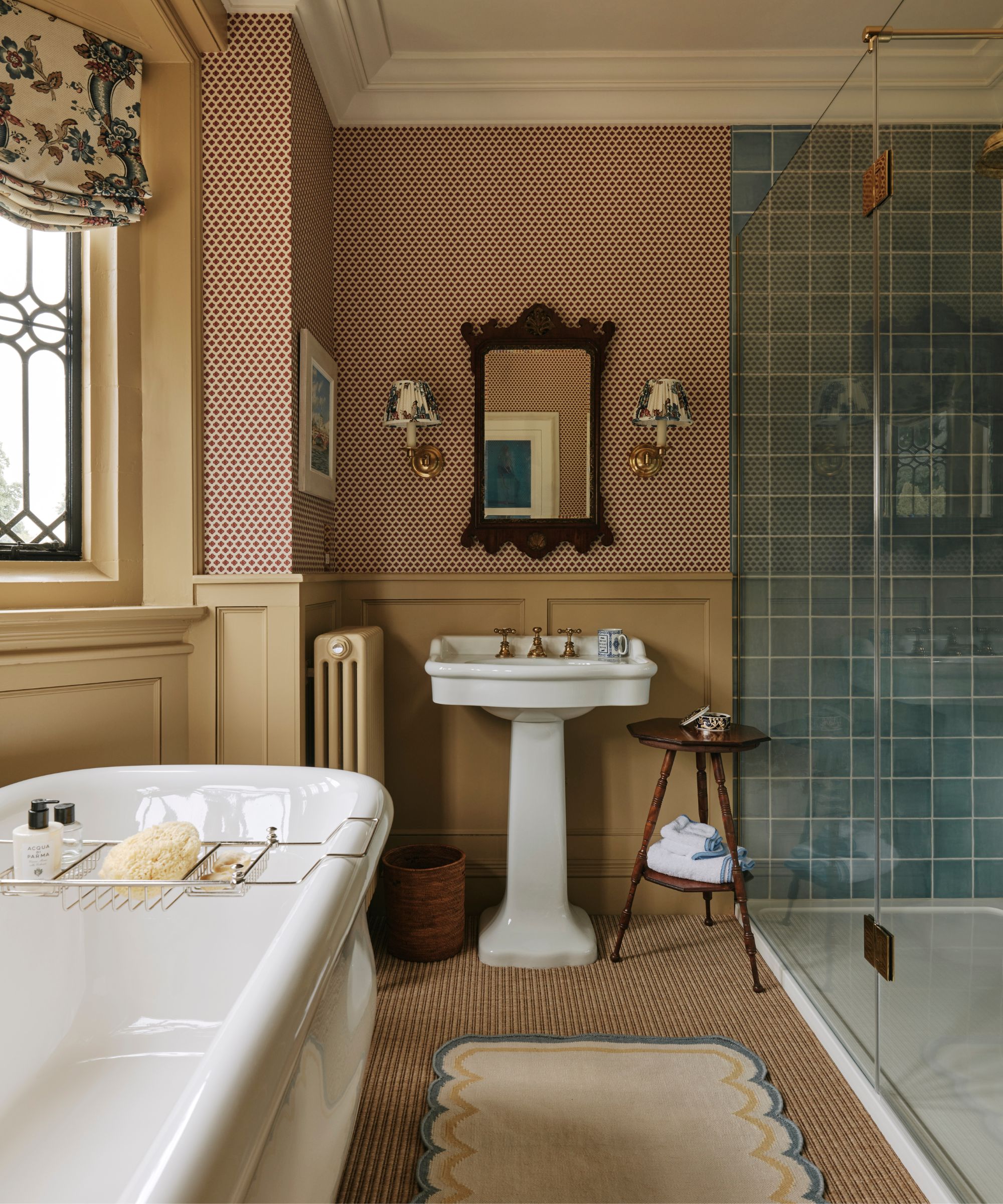 Bathroom with butter yellow panelling and window frame, polka dot wallpaper, blue tiled shower, jute carpet, pedestal sink and bath tub