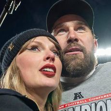 Taylor Swift wearing a black winter hat and hoodie hugging Travice Kelce on a football field with a light behind them