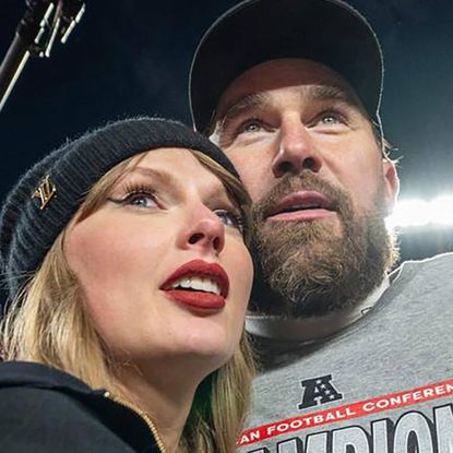 Taylor Swift wearing a black winter hat and hoodie hugging Travice Kelce on a football field with a light behind them