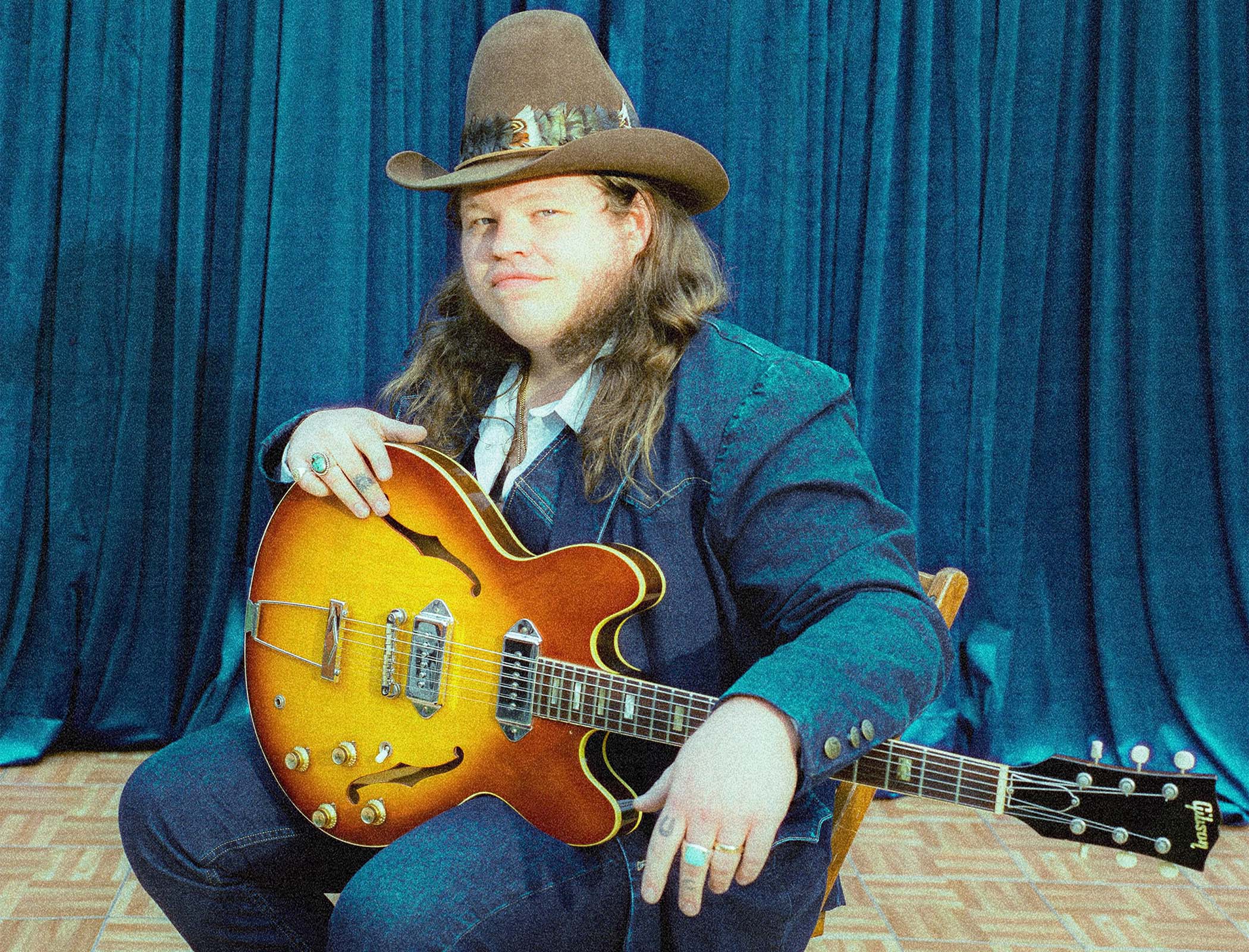 Marcus King photographed in front of blue velvet drapes, wearing a brown cowboy hat and seated with a Gibson ES-330