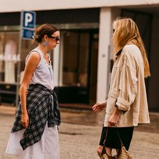 Two women are chatting wearing cute pics. One is wearing a dress over khaki trousers wit a plaid shirt around the waist. The other is wearing a barn jacket, pencil skirt, and mules. 