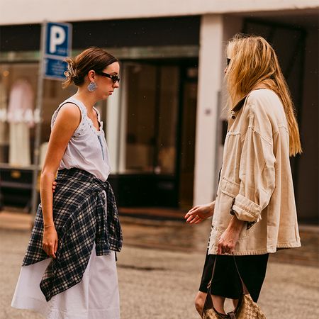 Two women are chatting wearing cute pics. One is wearing a dress over khaki trousers wit a plaid shirt around the waist. The other is wearing a barn jacket, pencil skirt, and mules. 