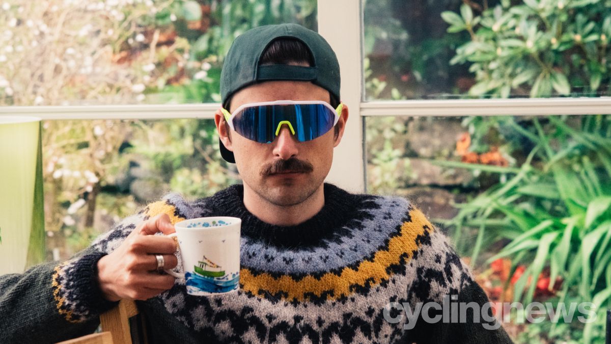 A white man in a backwards cap and a colourful jumper holds a mug of tea while wearing brightly coloured sunglasses