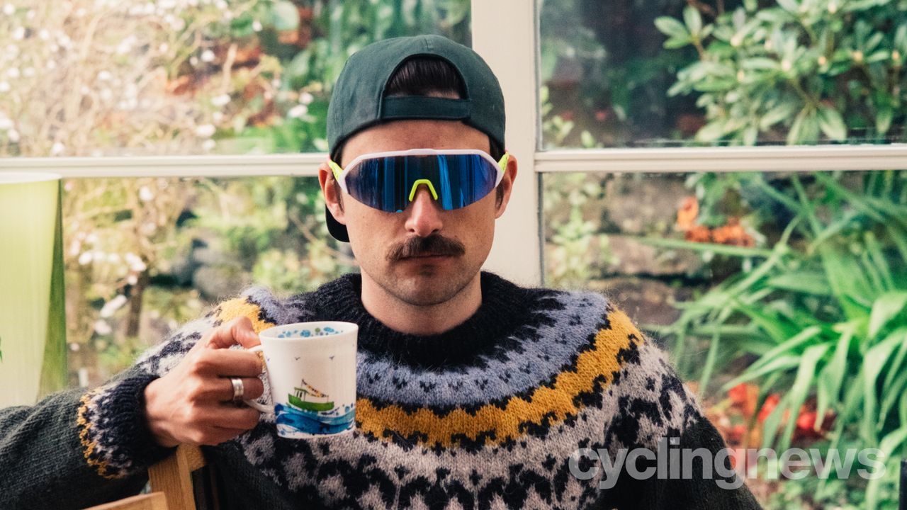 A white man in a backwards cap and a colourful jumper holds a mug of tea while wearing brightly coloured sunglasses