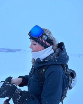 Princess Ingrid Alexandra smiling while wearing a blue coat and white scarf with goggles on her head putting on gloves in the snow