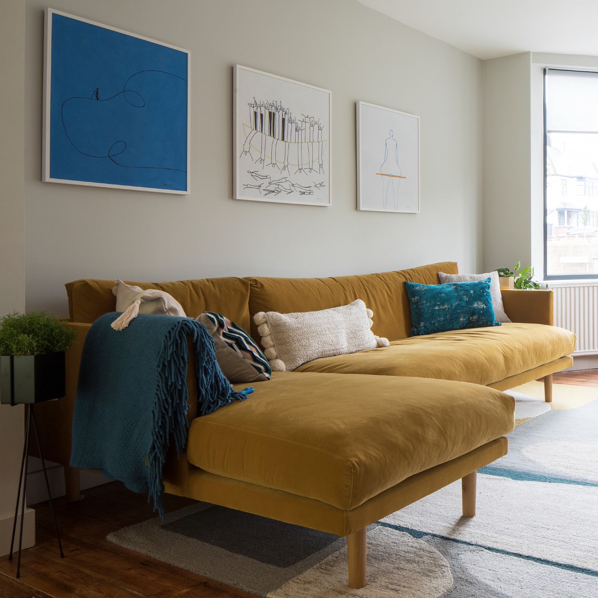 A living room with a mustard yellow velvet sofa and three artworks hung on the wall above it