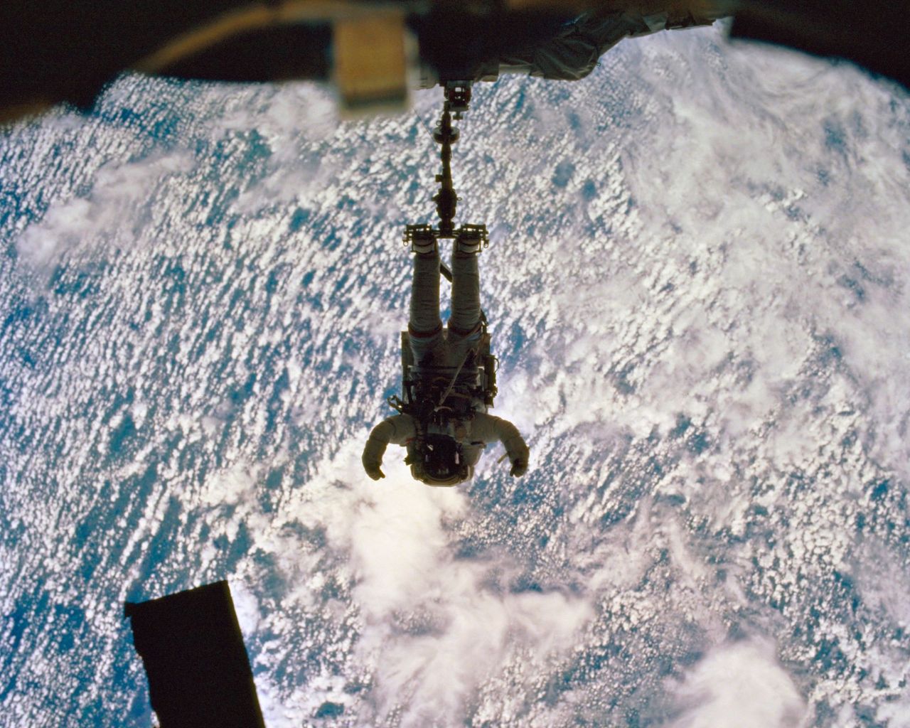 Chris Hadfield in full space suit and gear floating in space, attached by his feet to a harness and anchor, with the cloud-covered earth below him