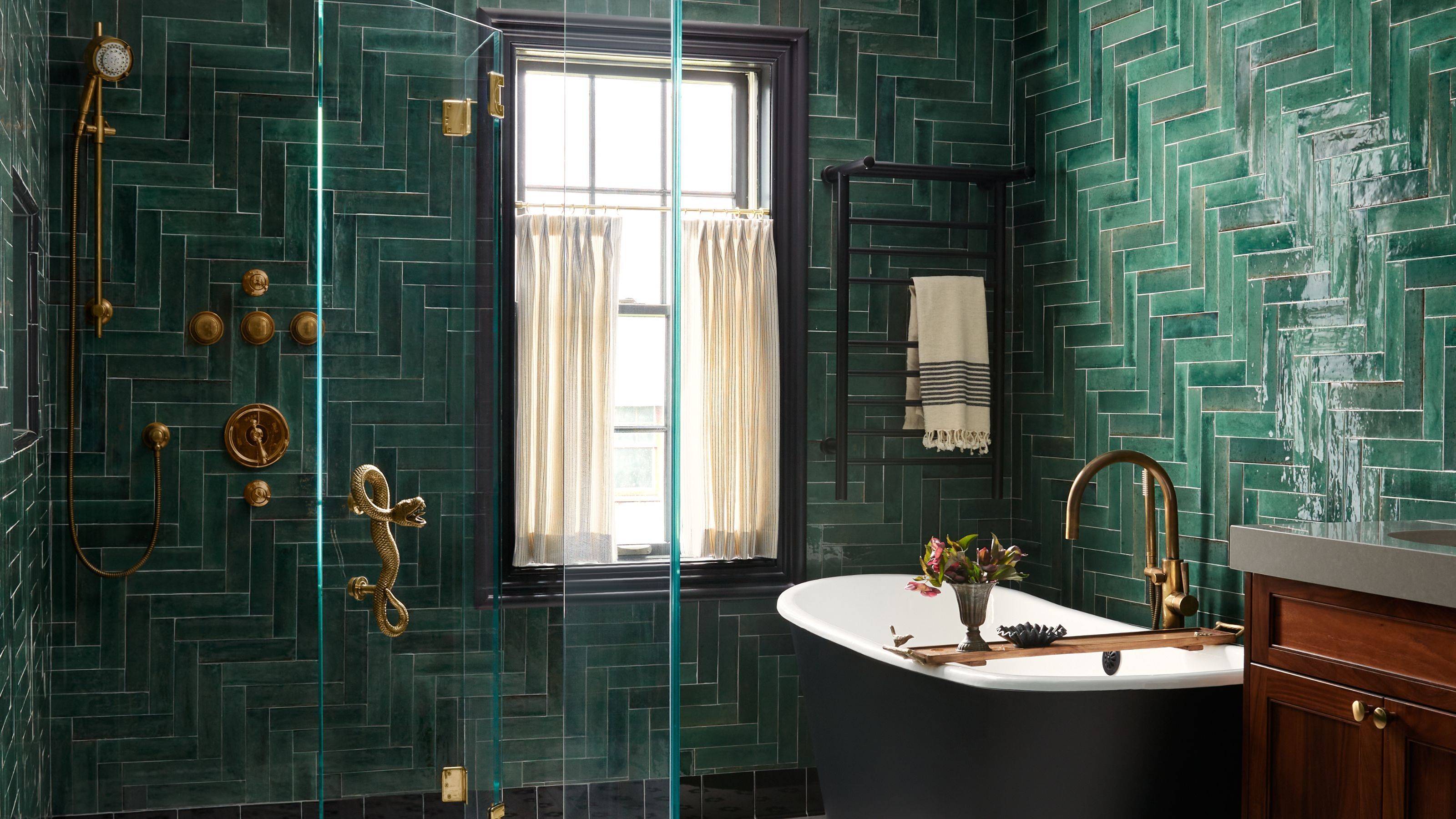 A green bathroom with dark to medium metro tiles in a pattern formation with brass details throughout, a freestanding bathtub, and a window with cafe curtains