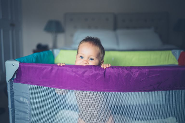 A happy little baby is standing in his travel cot and is looking over the side - the best travel cot buying guide