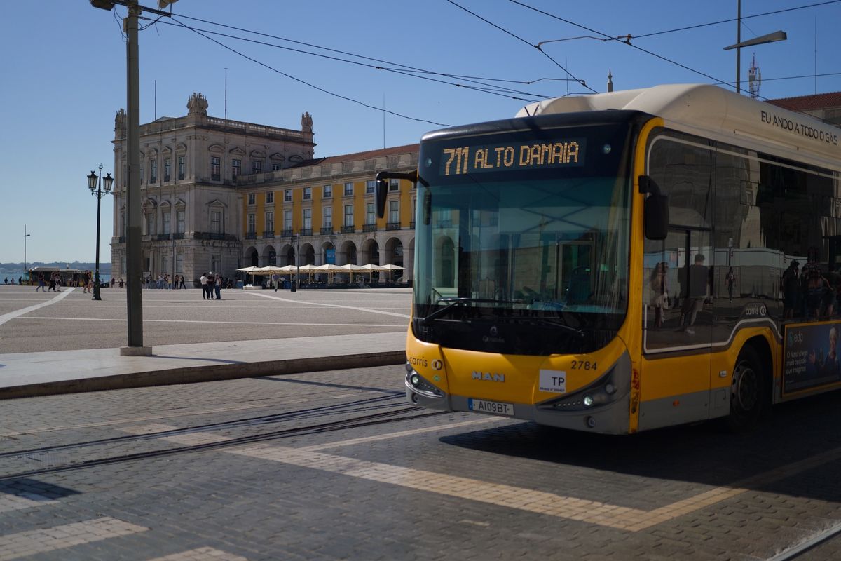 Yellow city bus crossing Pra&ccedil;a do Com&eacute;rcio in Lisbon, with historic buildings and overhead tram wires behind.