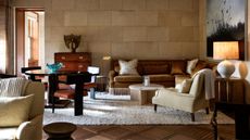 Warmly lit living room featuring stone block walls, a cognac velvet sectional, plush cream armchair, low marble coffee table, and dark wood dining chairs.