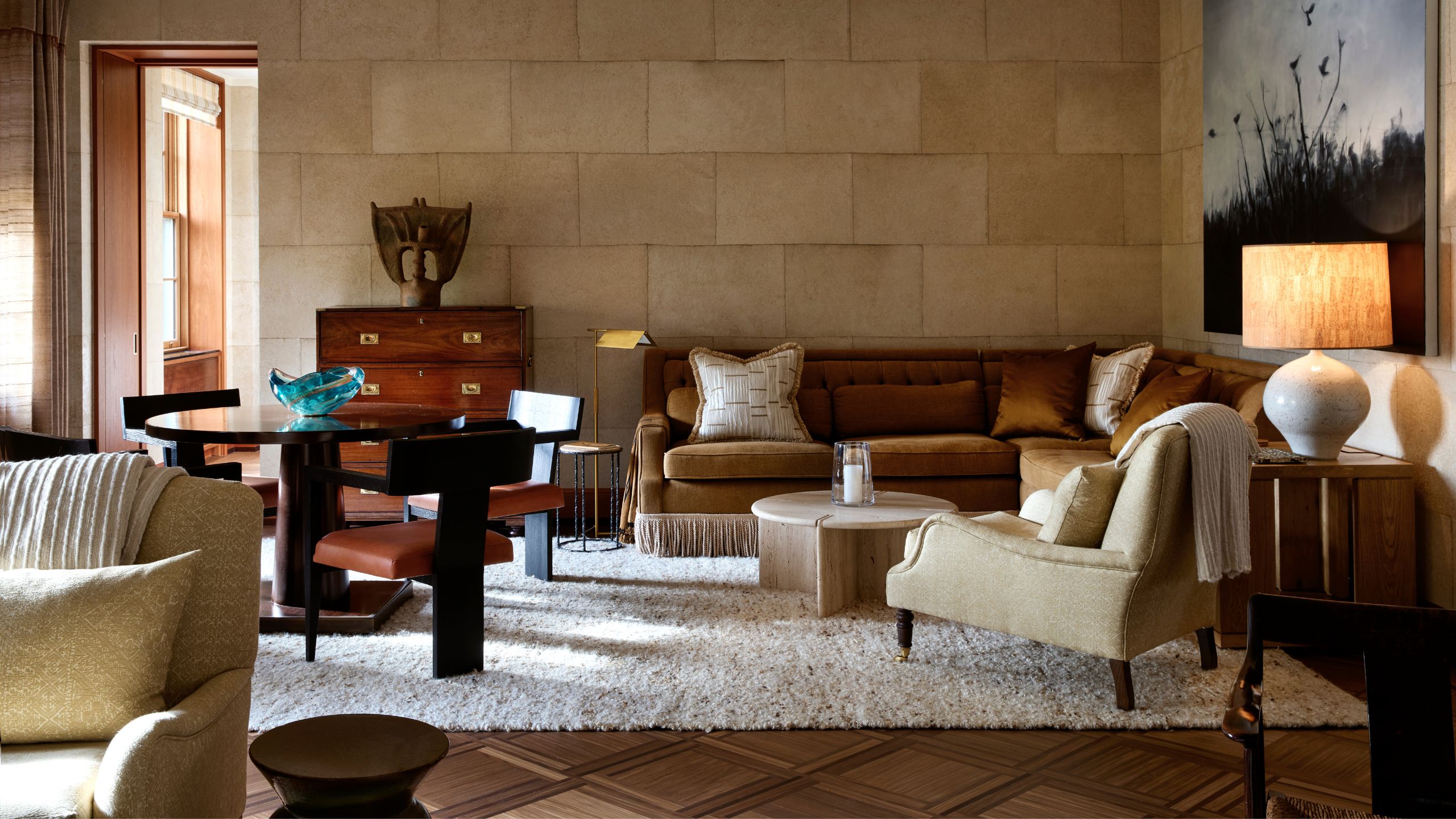 Warmly lit living room featuring stone block walls, a cognac velvet sectional, plush cream armchair, low marble coffee table, and dark wood dining chairs.