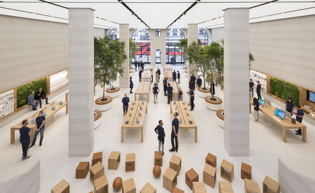 Interior view from an upper level of Apple&#039;s London flagship store featuring light coloured flooring, pillars, long wooden display tables with products on top, arched floor-to-ceiling windows, greenery on the walls and trees in round planters. There are multiple people inside the store