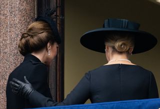 The back of Kate Middleton and Duchess Sophie, both wearing black dresses and hats, with Sophie putting her black gloved hand on Kate's shoulder