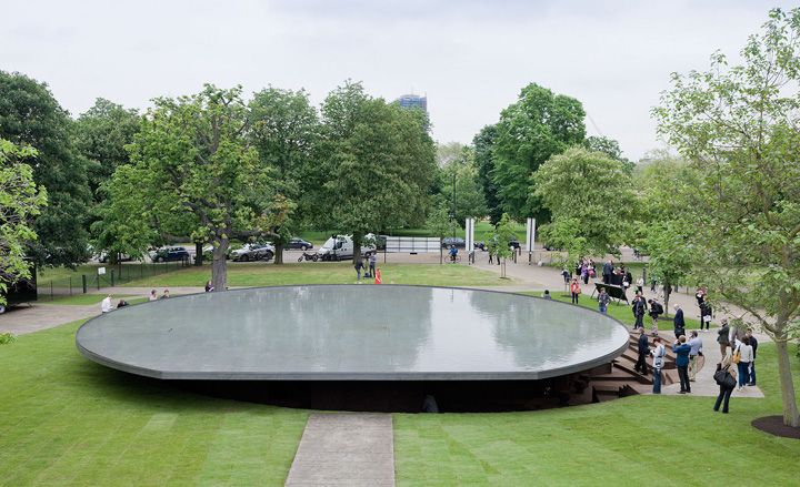 Serpentine gallery roof which holds a small pool of water surrounded with green grass, trees and people