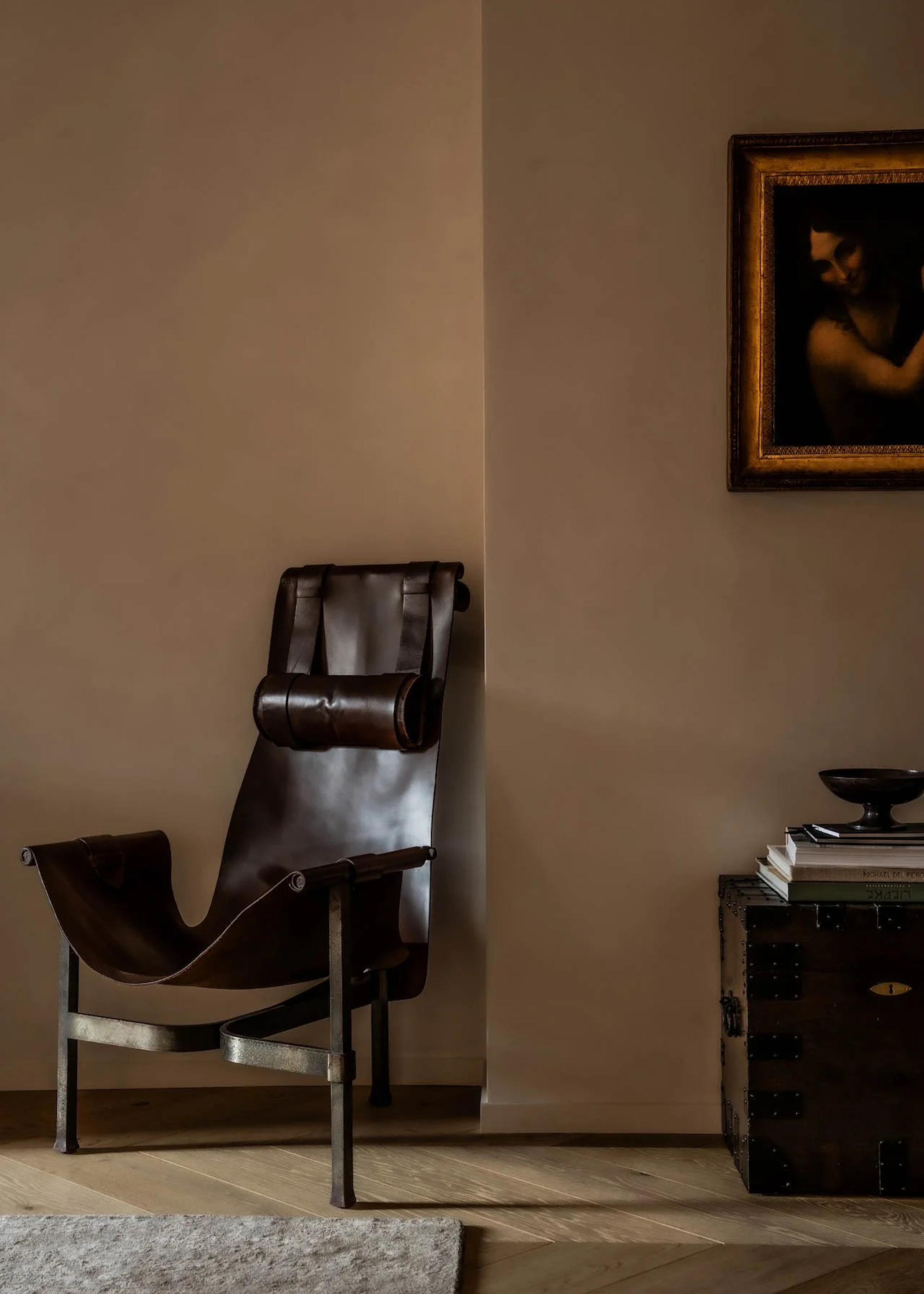 A living room with brown-beige walls, a leather antique seat, a framed painting, and a vintage trunk with a stack of books and a footed bowl