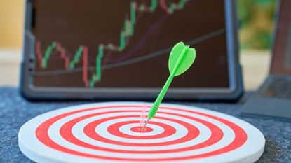 a green dart in the middle of a bullseye placed in front of a laptop with a blurred stock chart on the screen