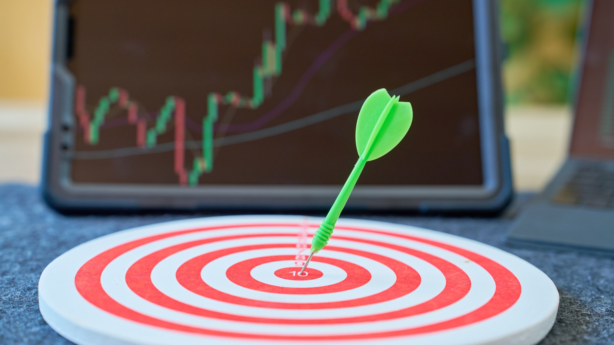 a green dart in the middle of a bullseye placed in front of a laptop with a blurred stock chart on the screen
