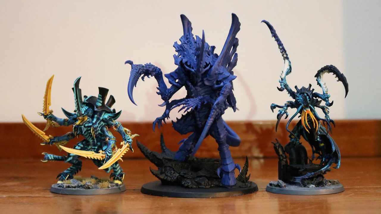 A size comparison between the Tyranid Norn Emissary and other miniatures, all sat on a wooden table