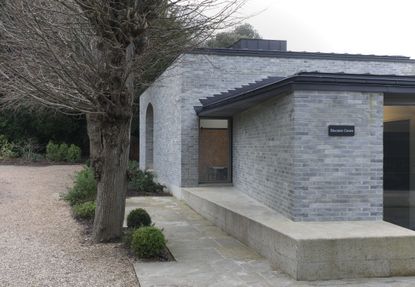 The facade of the education center is made out of gray bricks with a black roof. Next to the center are trees and foliage.