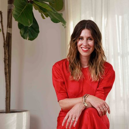 Colleen Bordeaux smiles while seated in a red top beside a large leafy plant and sheer white curtains.