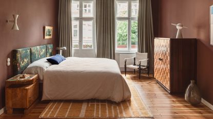 bedroom painted in warm clay color, tapestry headboard, natural linen bedding, timber floors, timber side table, marquetry cabinet with lamp on top and glass vase on the floor beside