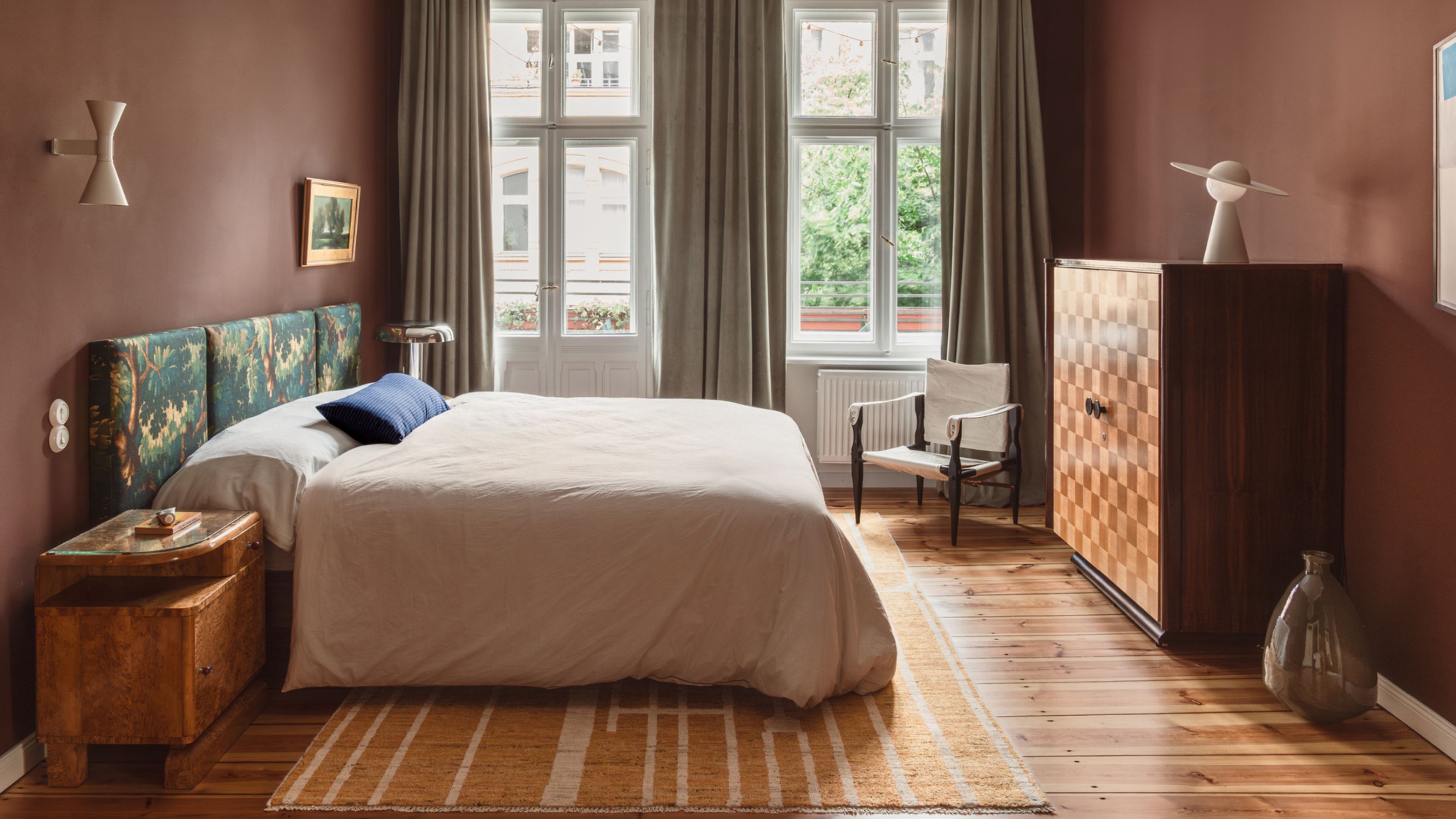 bedroom painted in warm clay color, tapestry headboard, natural linen bedding, timber floors, timber side table, marquetry cabinet with lamp on top and glass vase on the floor beside