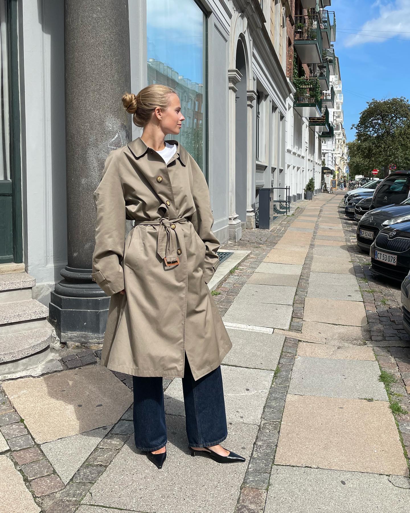 A criativa da moda dinamarquesa Clara Dyrhauge posa em uma calçada usando um casaco com cinto, camiseta branca, jeans de lavagem escura e slingbacks de salto de gatinho preto.