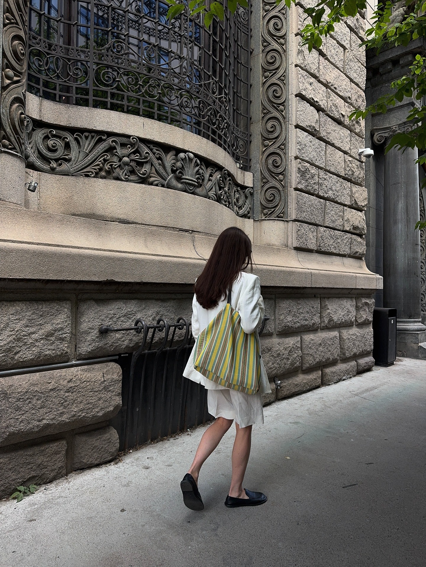 Woman wears white blazer, white trousers, loafers and carries striped The Row barn tote