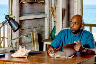 DI Mervin Wilson (Don Gilet) sits at the table in the beach hut where the Inspectors live in Death in Paradise. He is reading through some hard copy case files on the table in front of him, but looking suspiciously at Harry the Lizard, who is standing on the top of an anglepoise lamp on the far side of the desk.