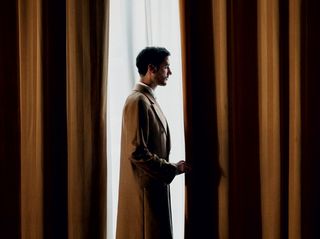 Man in brown trench coat looking out between brown curtains