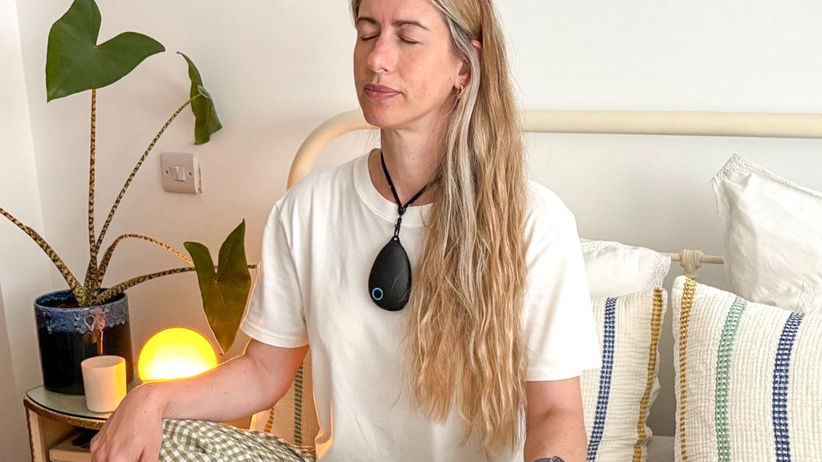 A blonde haired woman wearing a white t-shirt sitting crossed legged in bed with her eyes closed and her hands resting on her knees with a black, pebble shaped gadget around her neck
