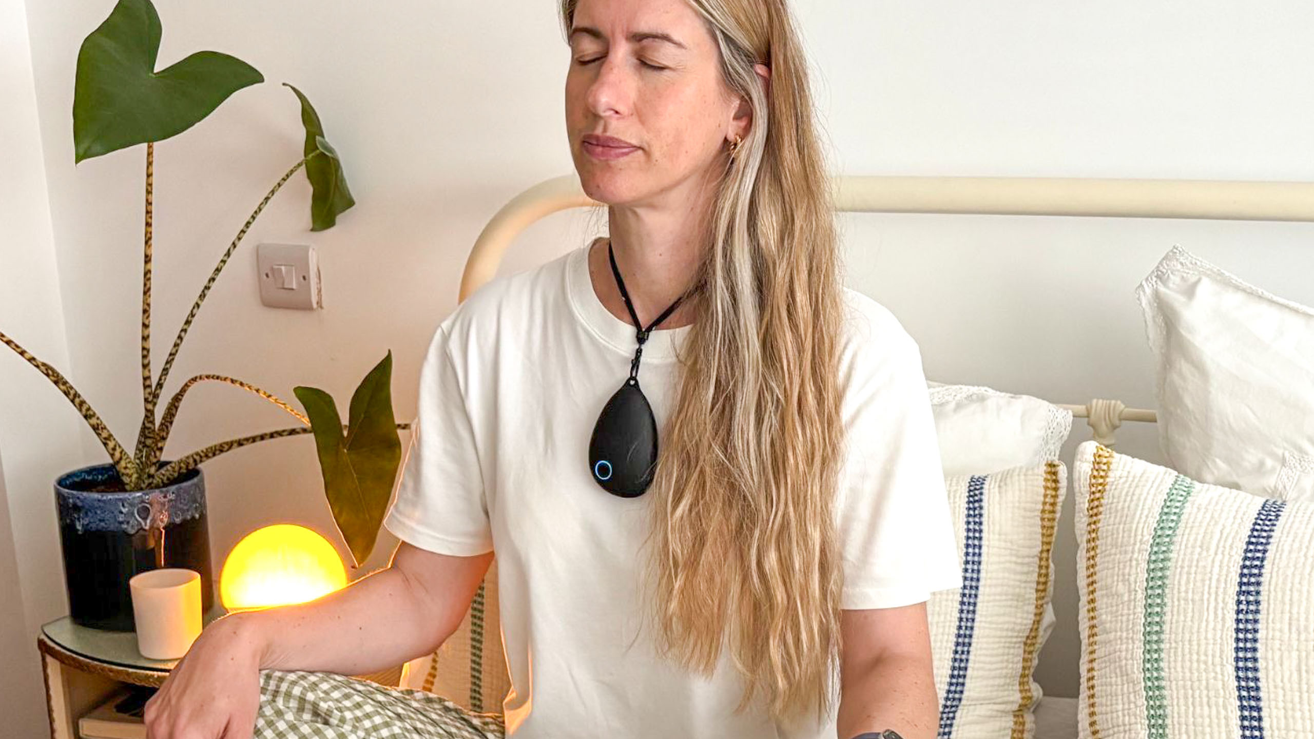 A blonde haired woman wearing a white t-shirt sitting crossed legged in bed with her eyes closed and her hands resting on her knees with a black, pebble shaped gadget around her neck