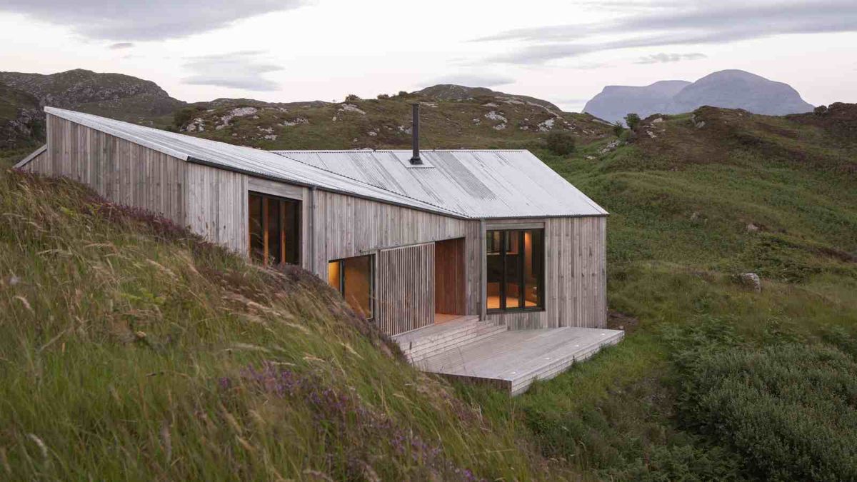 A contemporary timber-clad self build built into the slopes of a rural landscape in Scotland. The property is surrounded by long grasses and there are hills in the background. 