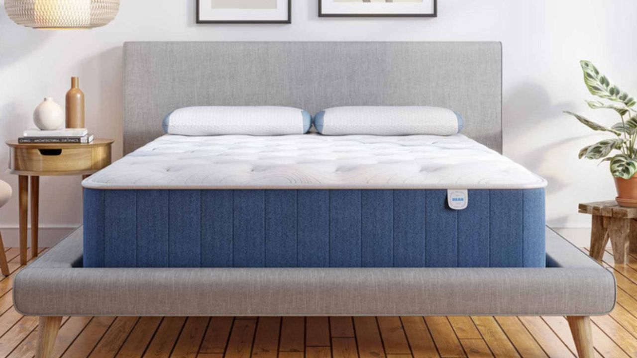 A plush mattress, the Bear Elite Hybrid Mattress, on a bed beside a nightstand against a white wall.